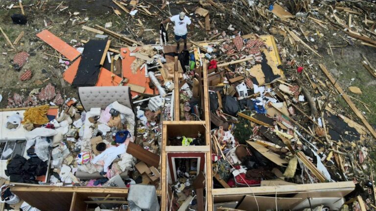 Un hombre, arriba en el centro, rescata una chaqueta mientras observa una casa dañada por un tornado, el domingo 26 de marzo de 2023, en Rolling Fork, Mississippi.