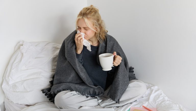 Portrait of woman catching a flu, sneezing, feeling sick, sitting on bed with laptop and working on remote while caught a cold